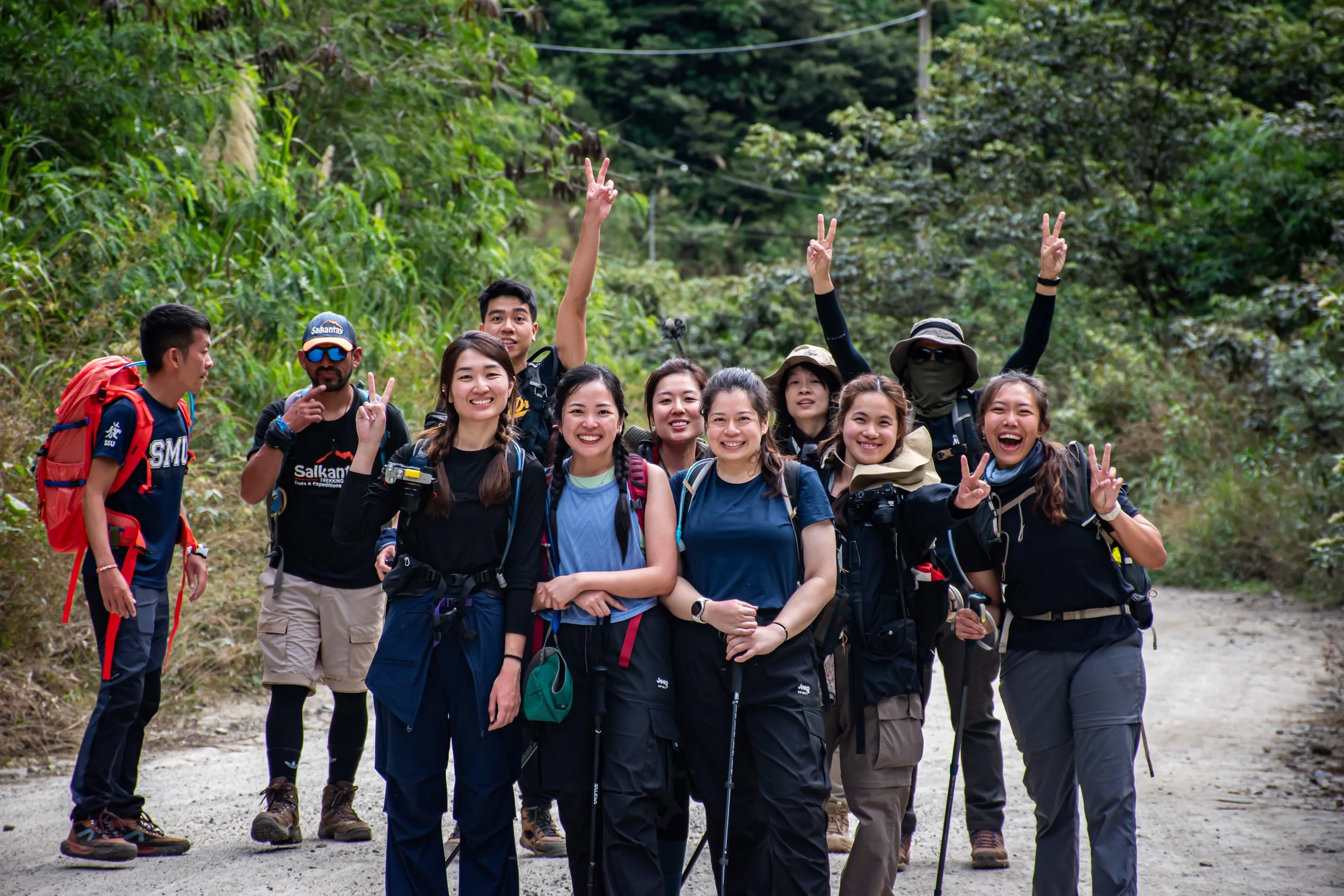 A group of ten hikers, smiling and posing for a photo on a forest trail. They are wearing hiking gear including backpacks, hats, and trekking poles, surrounded by green trees.