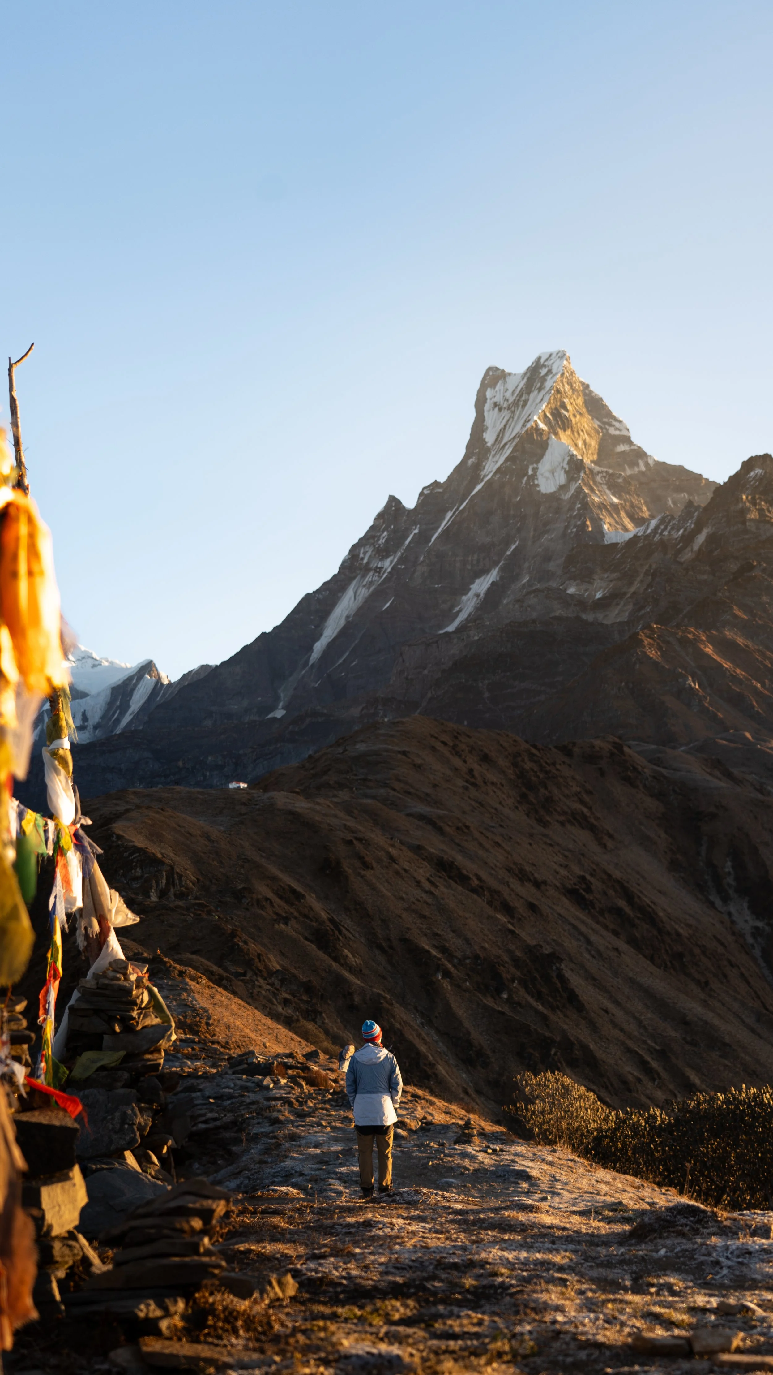 A person walking on a hiking trail in the mountains with prayer flags on the side, tall snow-capped peaks in the background, and a clear blue sky.