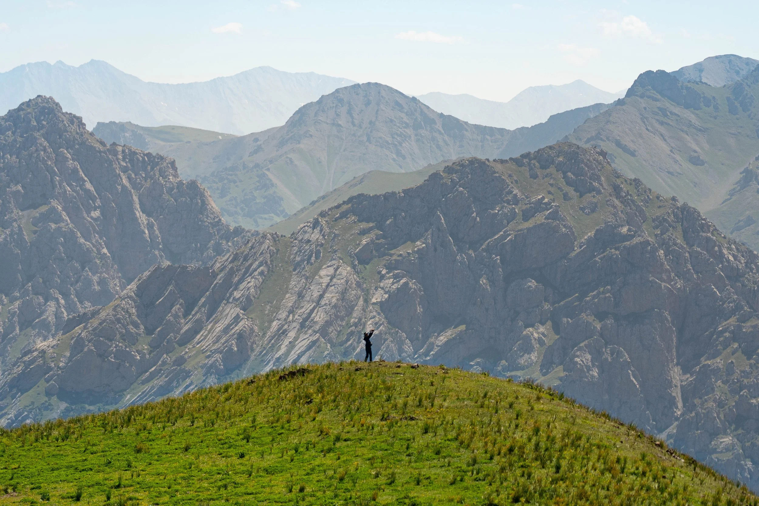 A person standing on a grassy hill at the base of a mountain range with rugged peaks, in a scenic outdoor landscape.