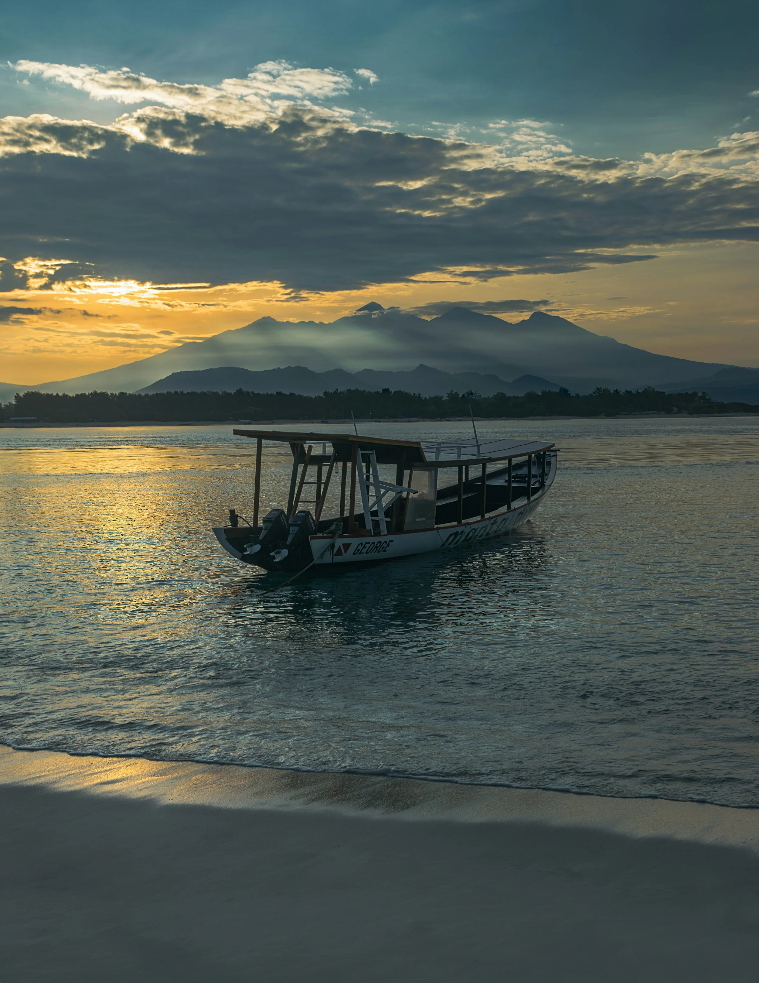 Gili island A boat floating on calm water at sunset with mountains in the background and partly cloudy sky.