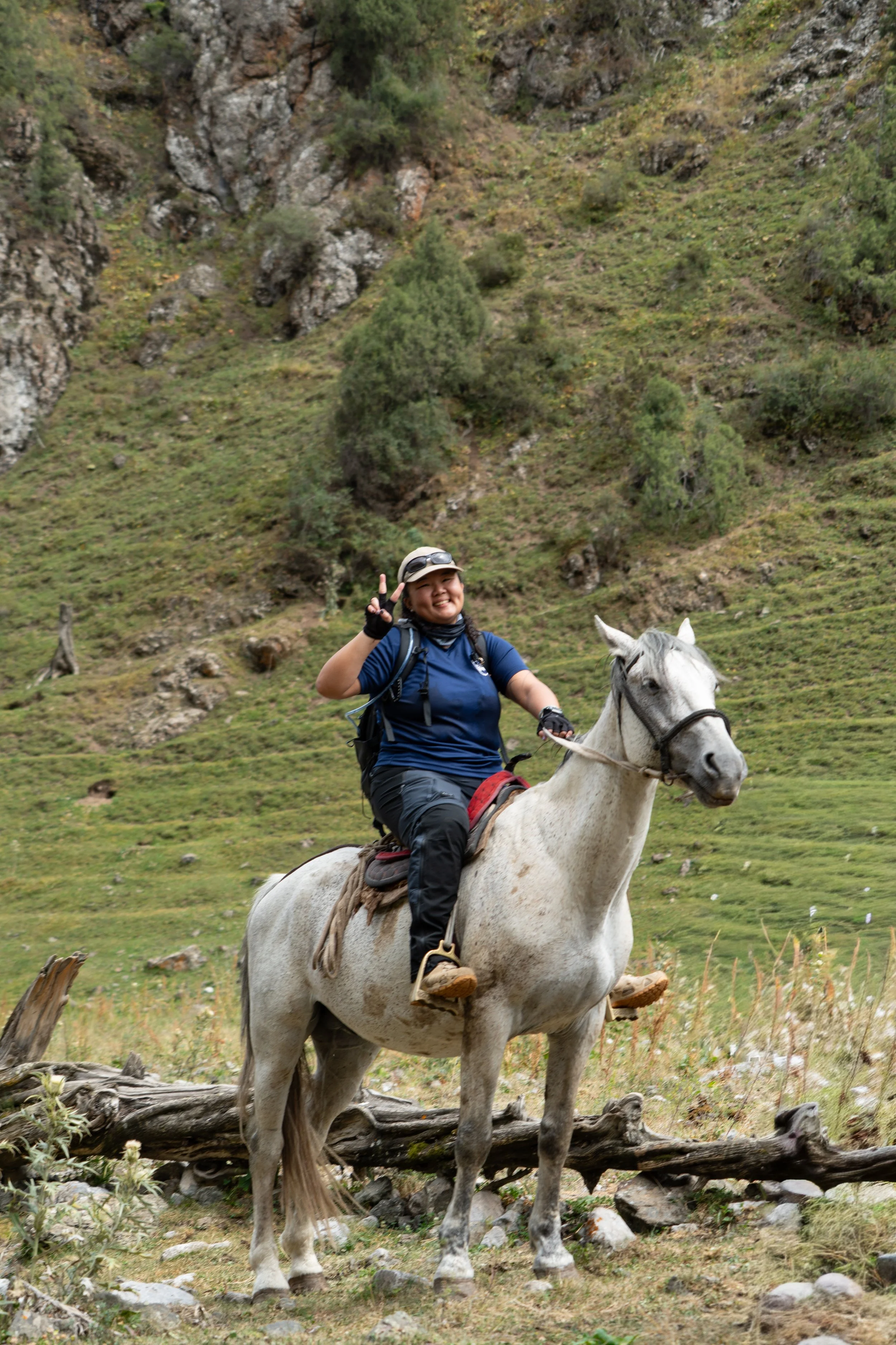 Kyrgyzstan Osh. A woman riding a white horse in a natural outdoor setting with green grass, rocks, and trees on a hillside, smiling and making a peace sign.