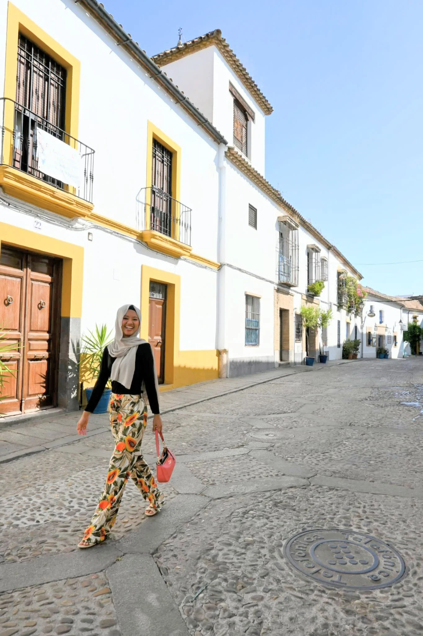 A smiling woman in floral pants and a black top, holding a pink purse, walking along a cobblestone street in a picturesque town with white buildings and balconies under a clear blue sky.
