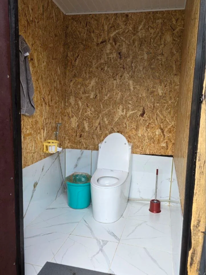 A small, unfinished bathroom with a white toilet, a blue trash bin, a yellow soap holder, and a red toilet brush. The walls are partially covered with particle board, and the floor is tiled with white marble-patterned tiles.