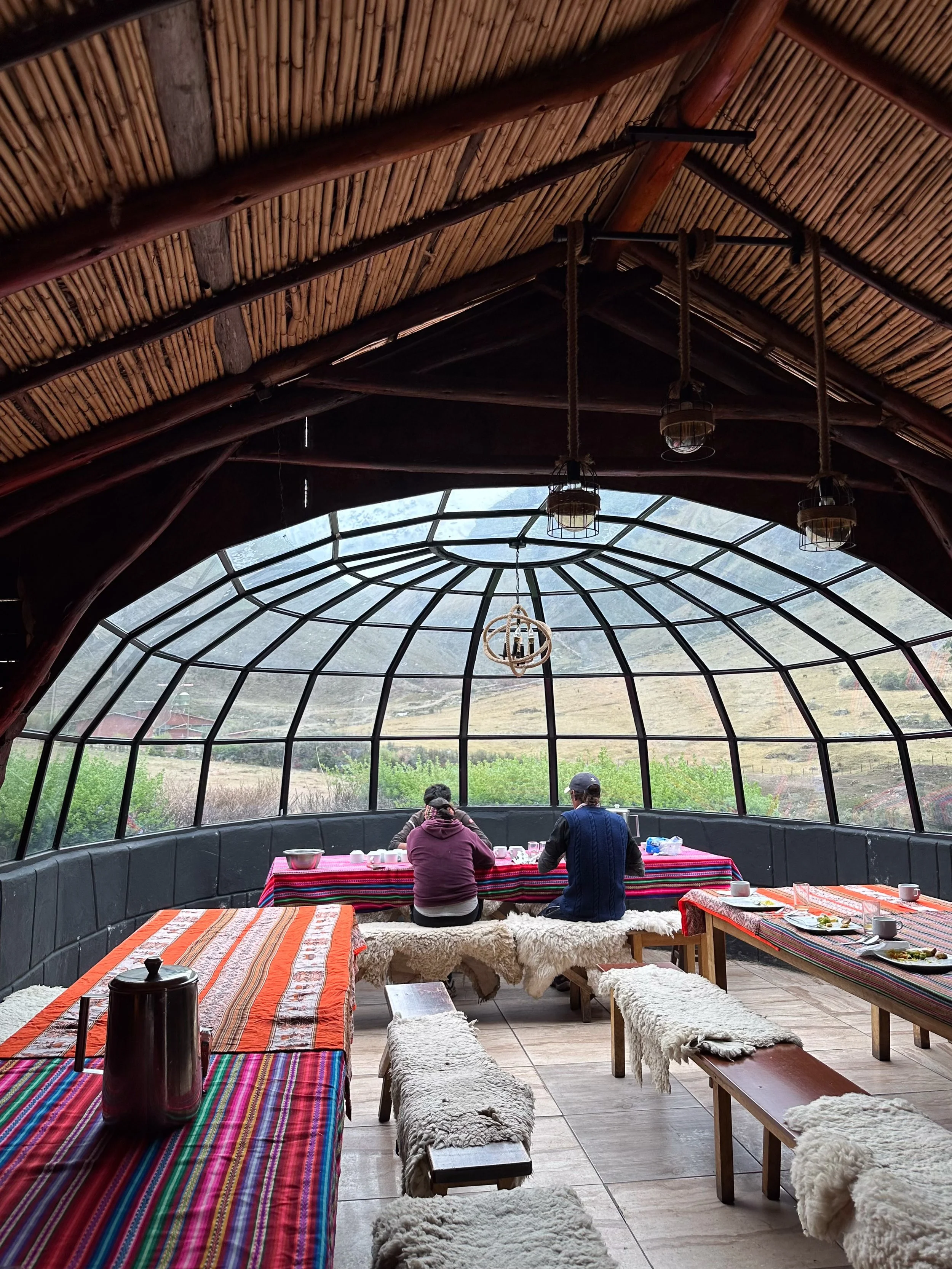 Peru, salkantay Trek glass domes. People sitting at a table inside a glass-roofed structure with mountain scenery outside, decorated with colorful striped tablecloths and sheepskin-covered benches.
