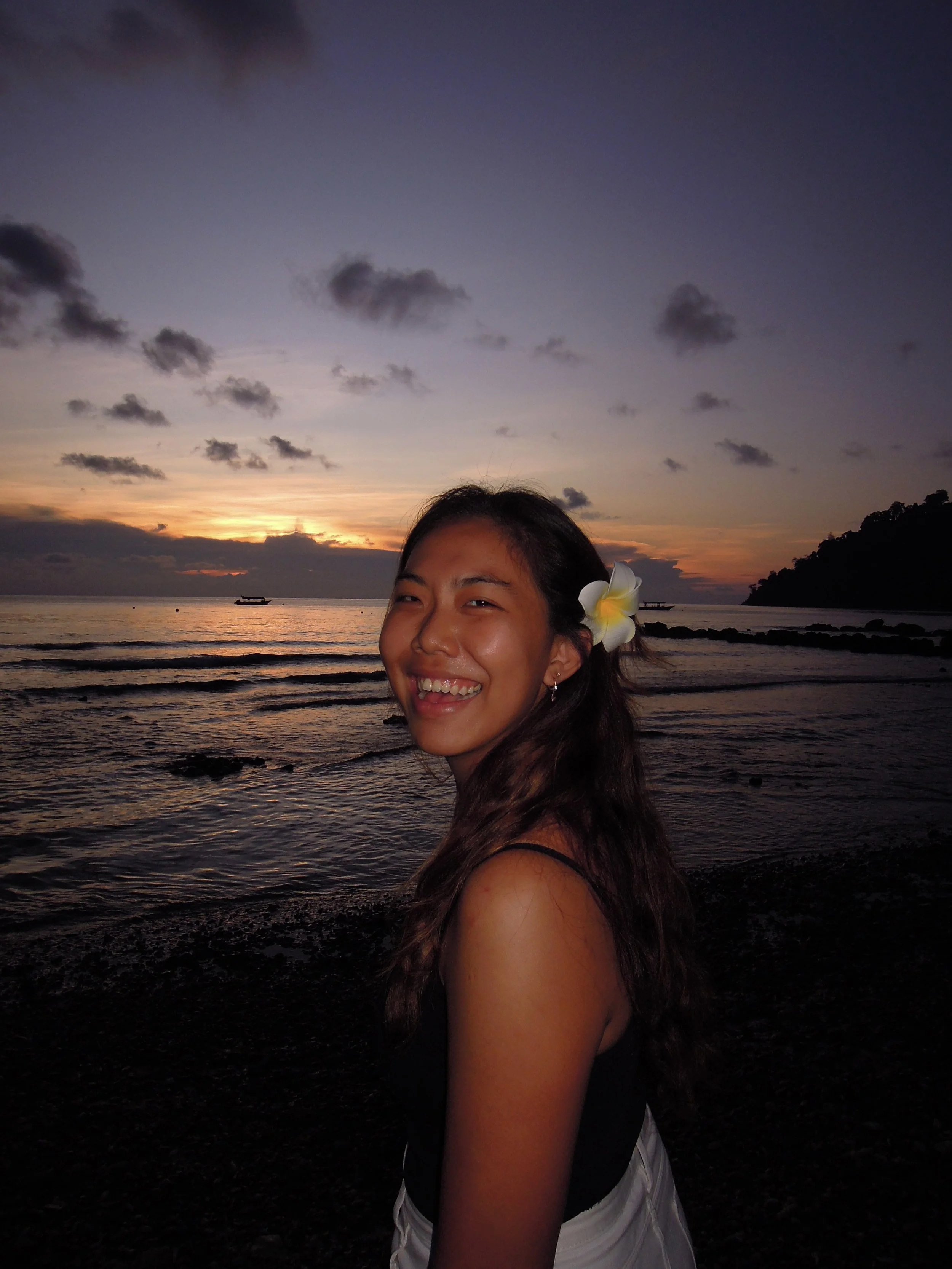 A young woman smiling on the beach at sunset with a flower in her hair.