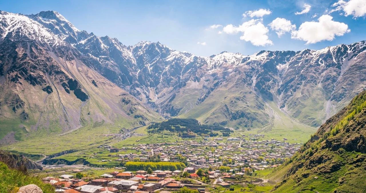 A panoramic view of a small town nestled in a valley surrounded by tall, snow-capped mountains with a partly cloudy sky.
