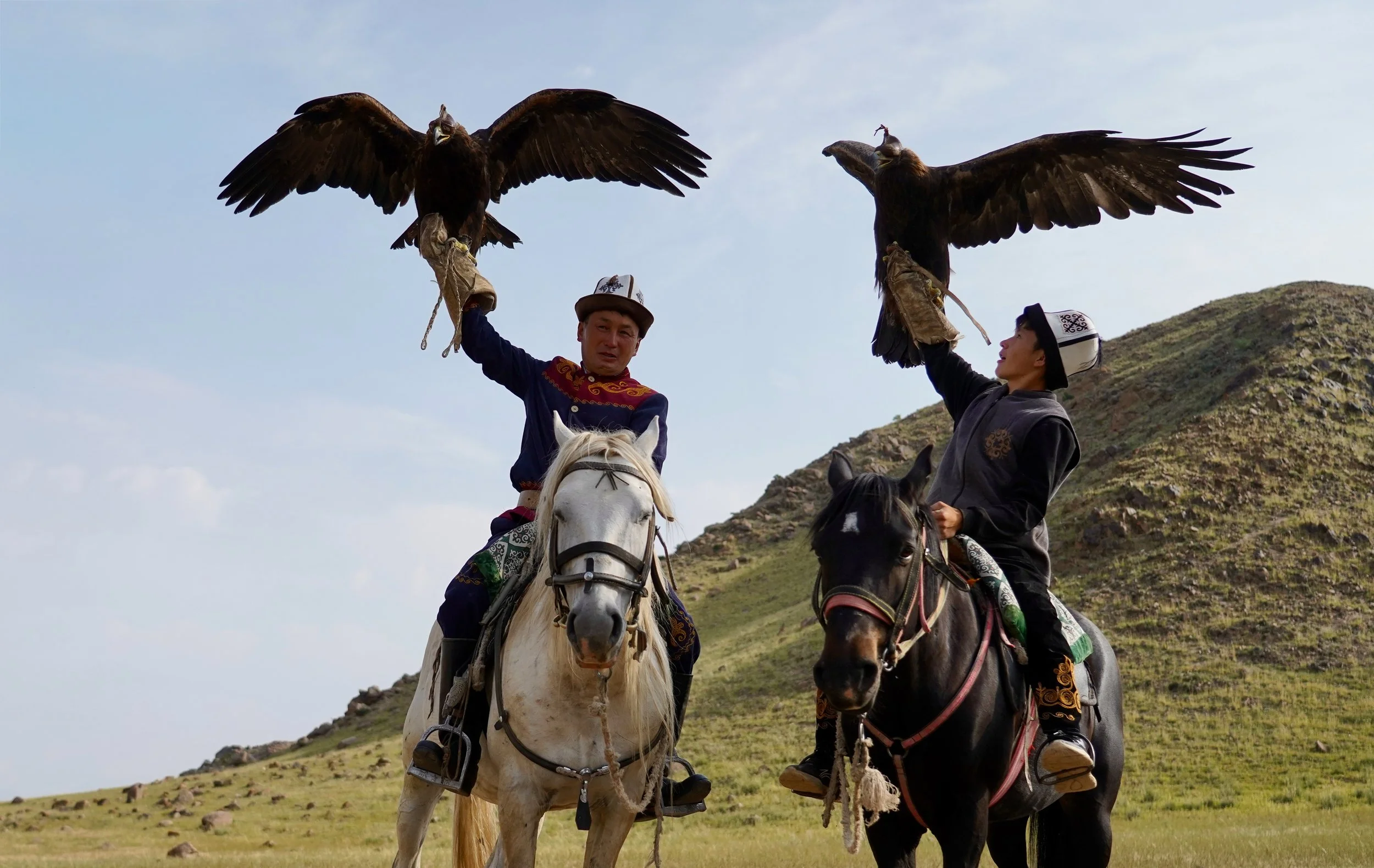 Two men on horseback holding up large eagles on their gloved hands in a grassy mountainous landscape.