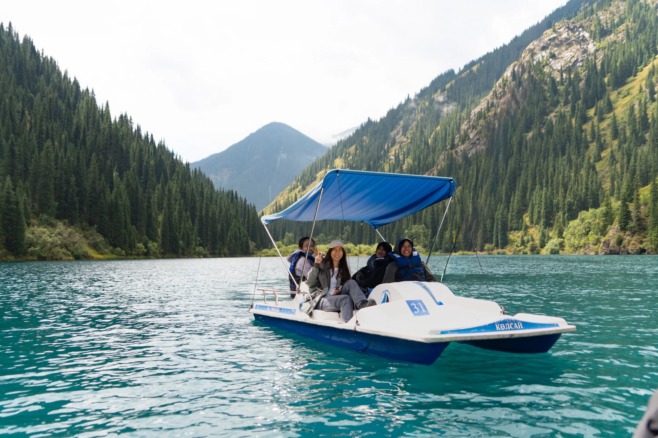 Kazakhstan Kolsay Lake Four people riding a boat on a mountain lake surrounded by dense evergreen trees and mountains.