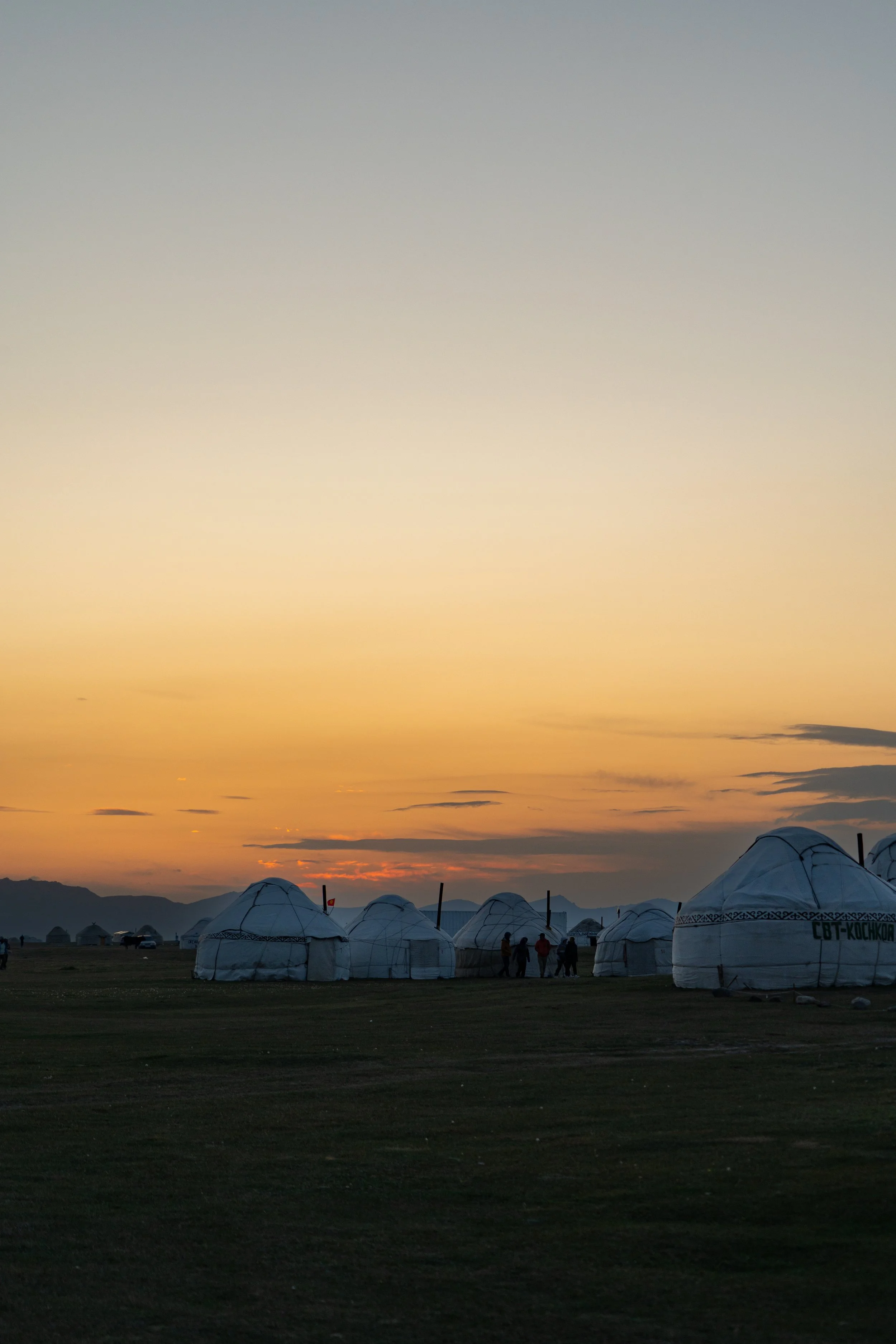 Kyrgyzstan. White traditional yurts on a grassy field at sunset with a few people standing near the yurts, mountains in the background, and a colorful sky.