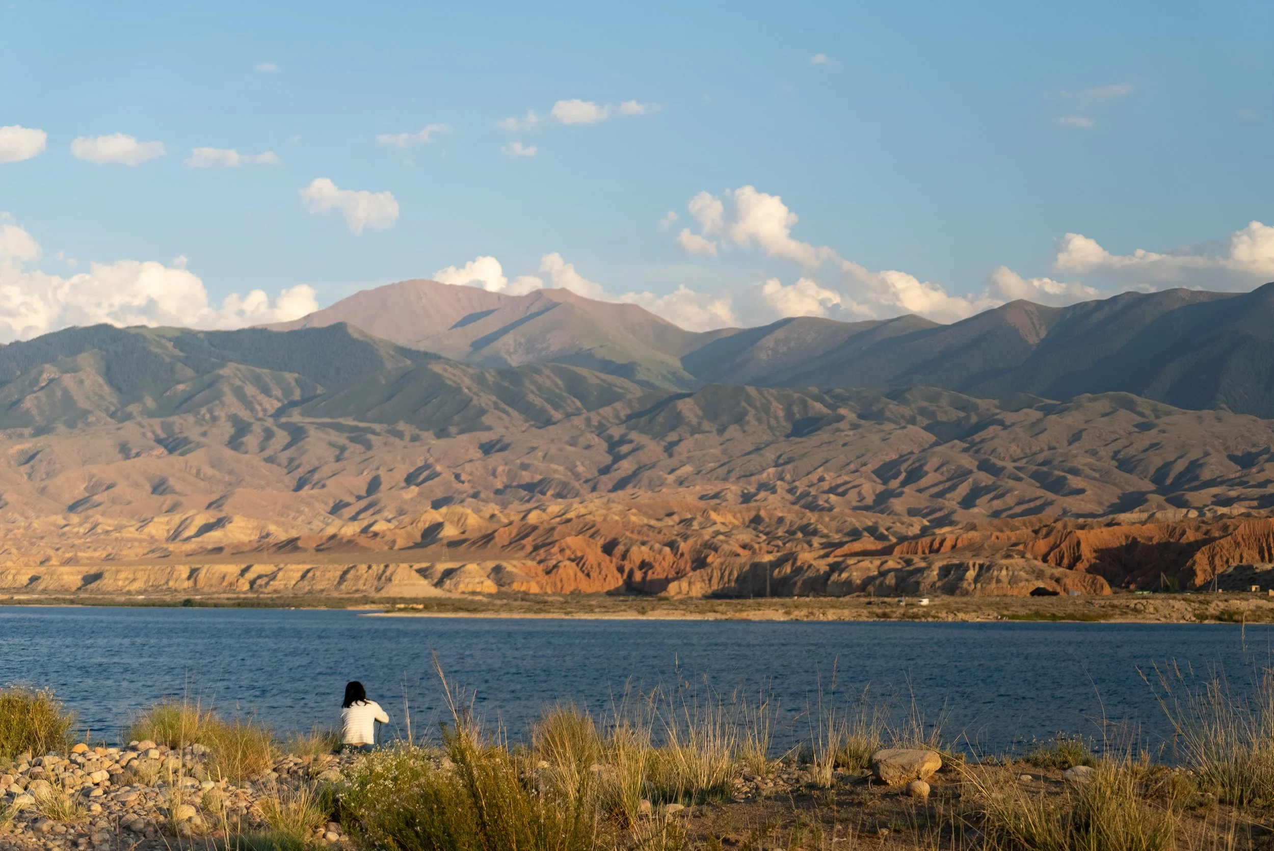Kazakhstan, Issyk kul Person sitting on rocky shore near a body of water, with mountains in the background under a partly cloudy sky.