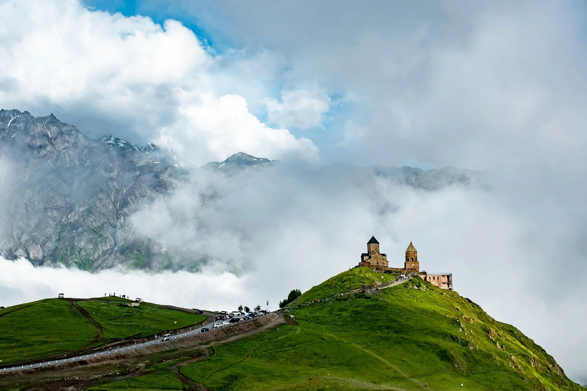 A church with two steeples situated on a grassy hilltop surrounded by clouds and mountains in the background.