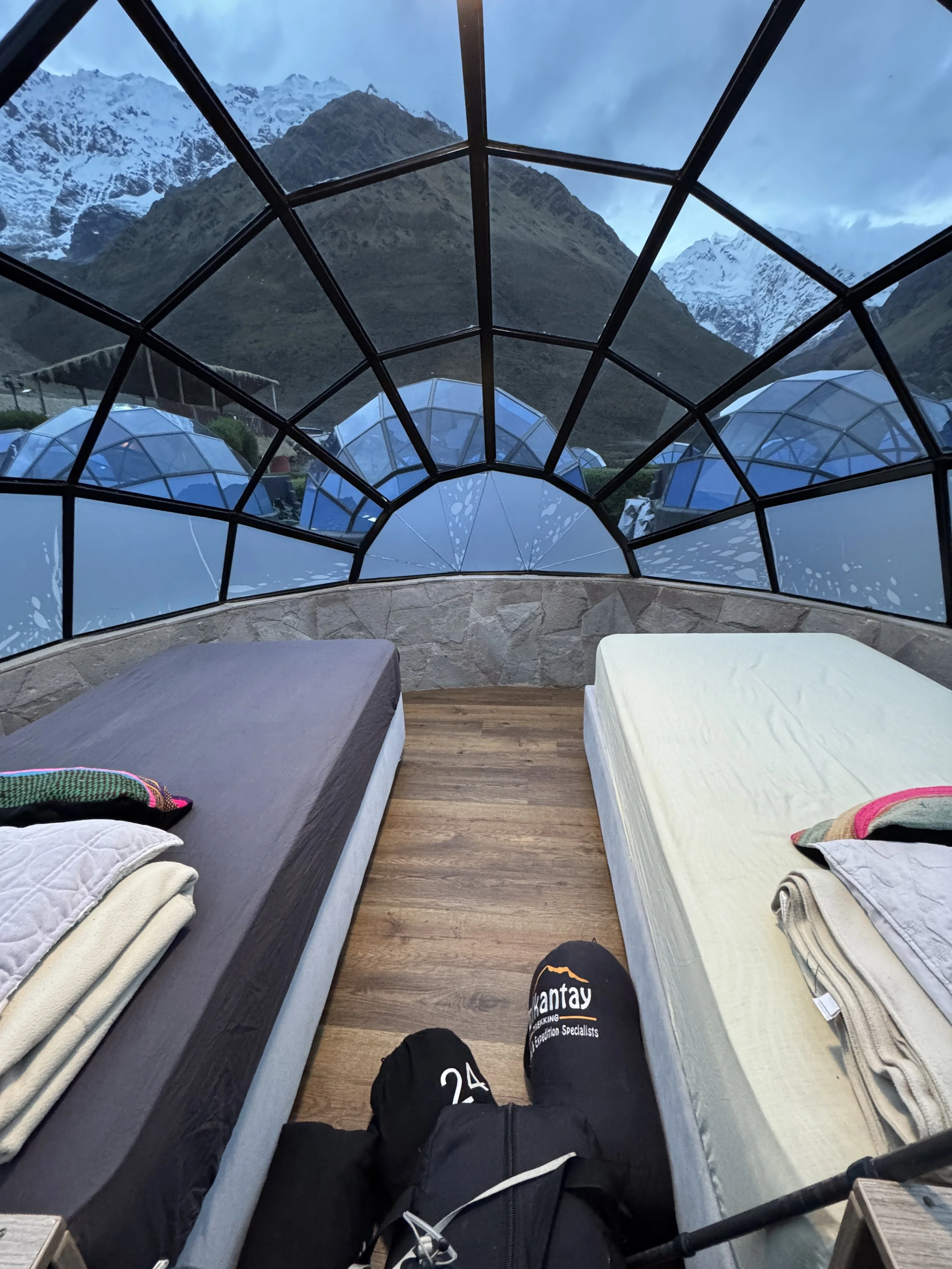 Two beds inside a glass-domed structure overlooking mountains with snow-capped peaks in the background.