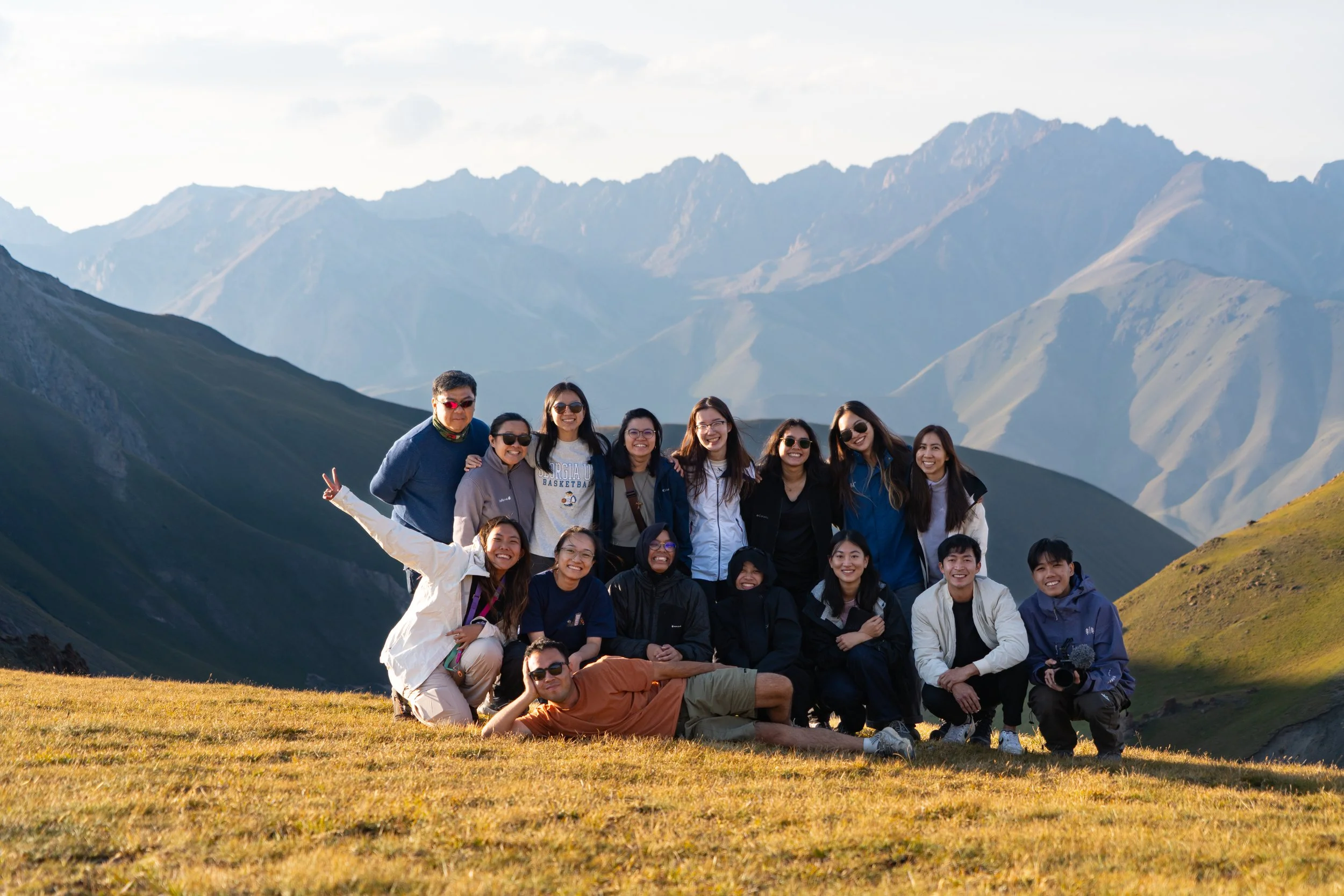 Group of people posing outdoors on a grassy hillside with mountains in the background during daytime.