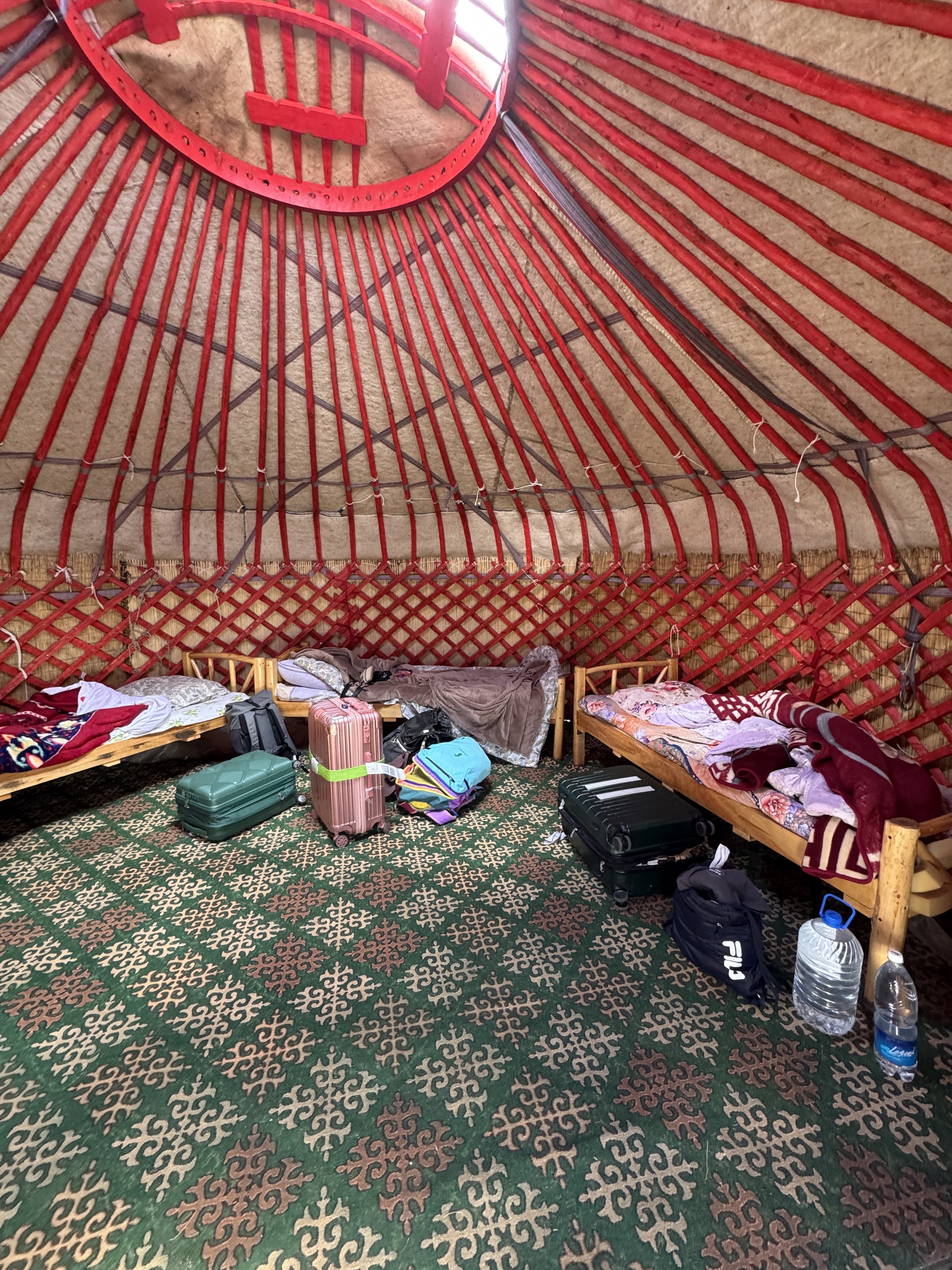 Inside a traditional yurt with wooden beds, scattered luggage, and water bottles on a patterned carpet.