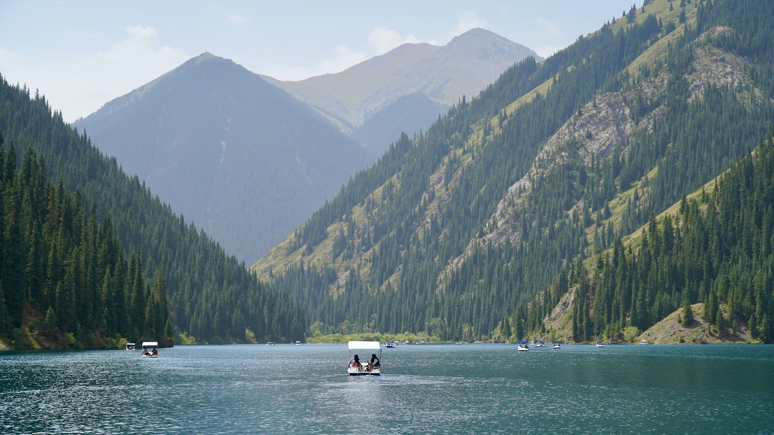 A lake surrounded by tall, green, forested mountains with a few boats on the water and a partly cloudy sky overhead.