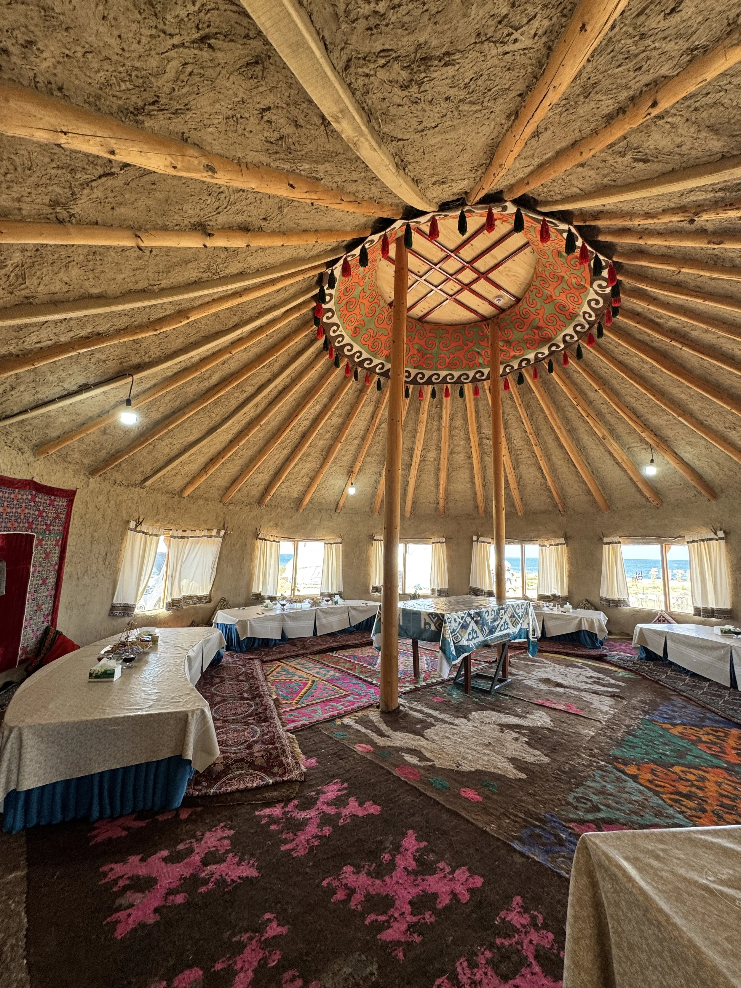 Interior of a round yurt with a high, thatched ceiling supported by wooden beams. Multiple tables with white tablecloths are set around the room, and several narrow windows with curtains let in natural light. The floor is decorated with colorful, patterned rugs.