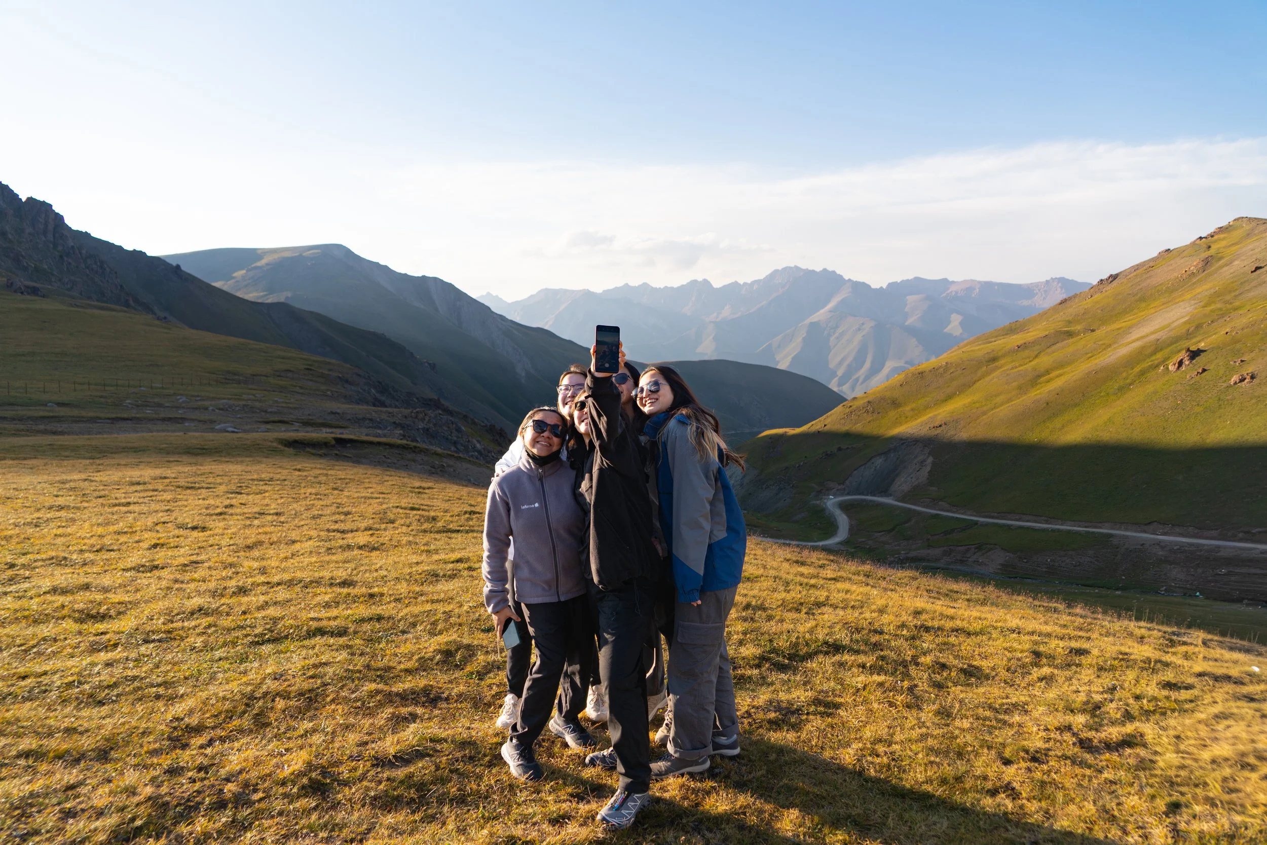 Group of four people taking a selfie on a grassy mountain landscape during sunset, with rolling hills and mountain peaks in the background.