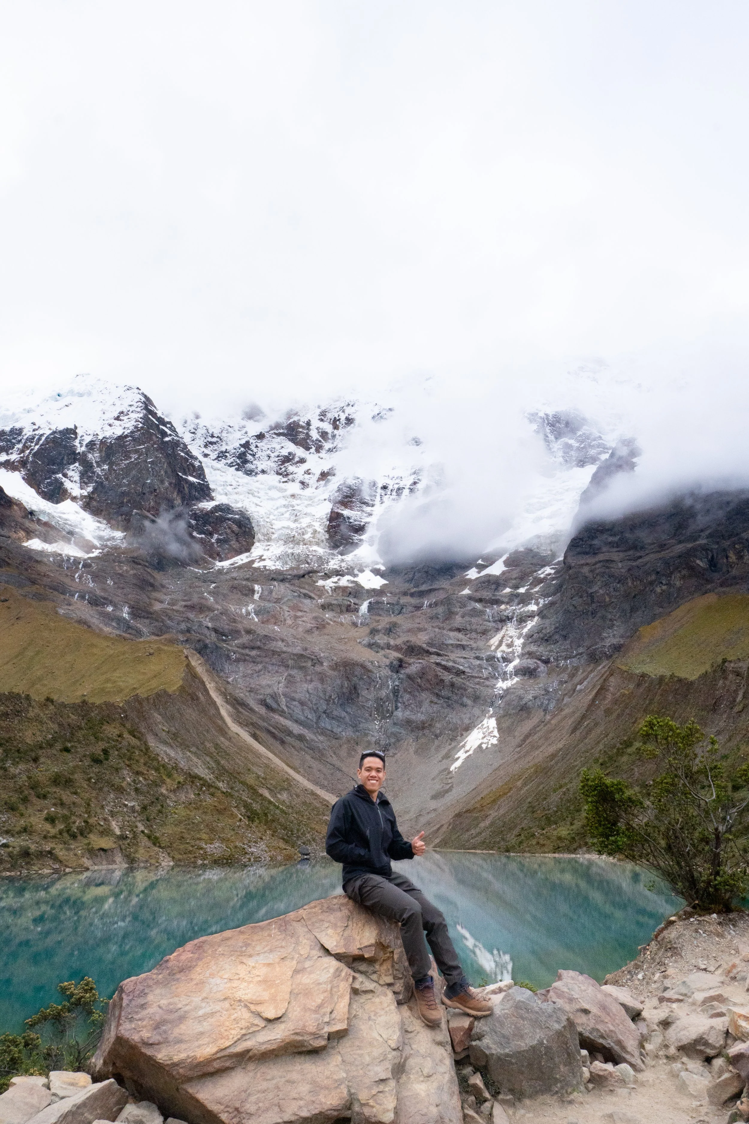 A person sitting on a large rock with a lake, mountain, and snow-covered peaks in the background.