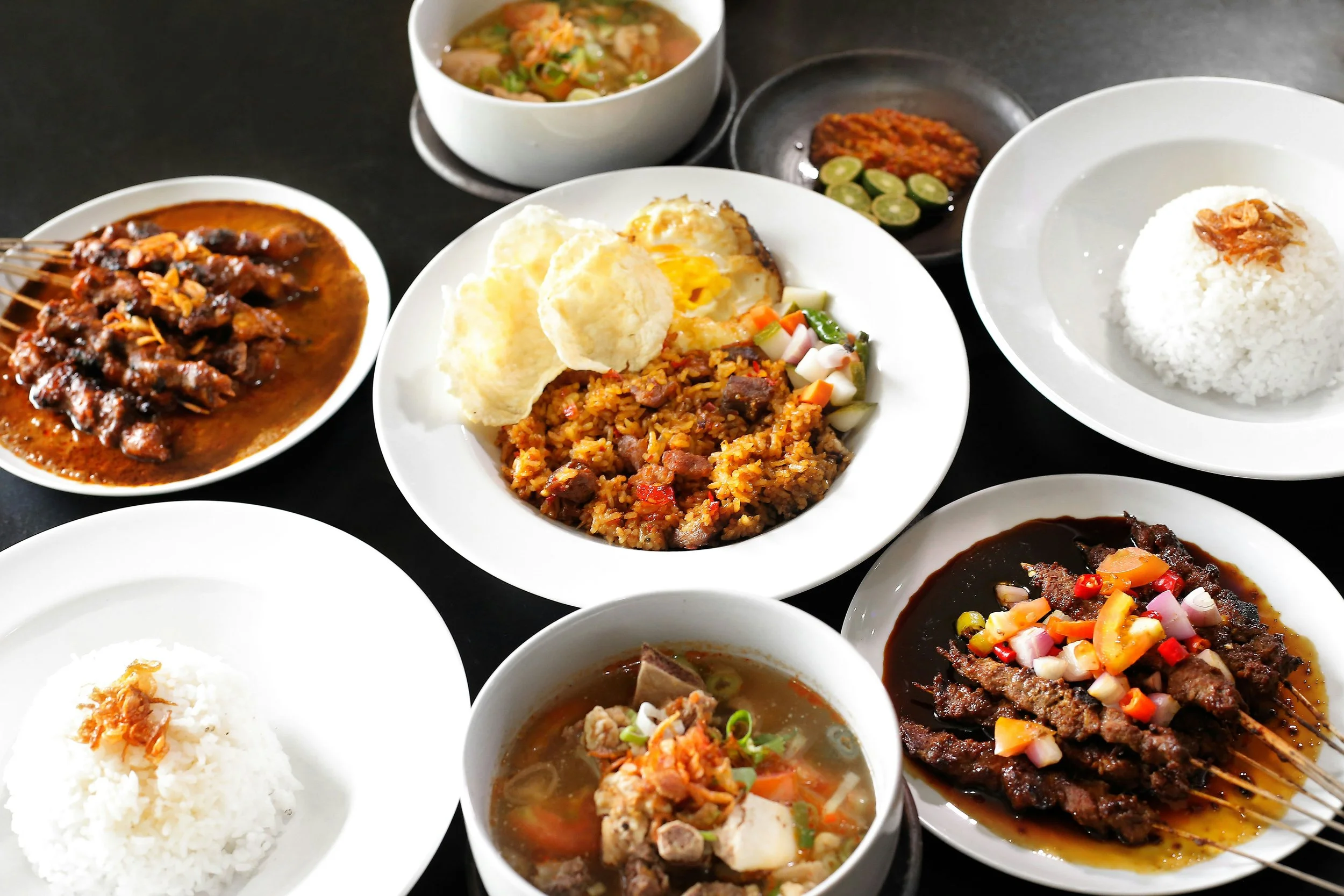 Assorted Indonesian dishes including rice, satay skewers, fried rice with vegetables, vegetable soup, and side condiments on black and white plates.