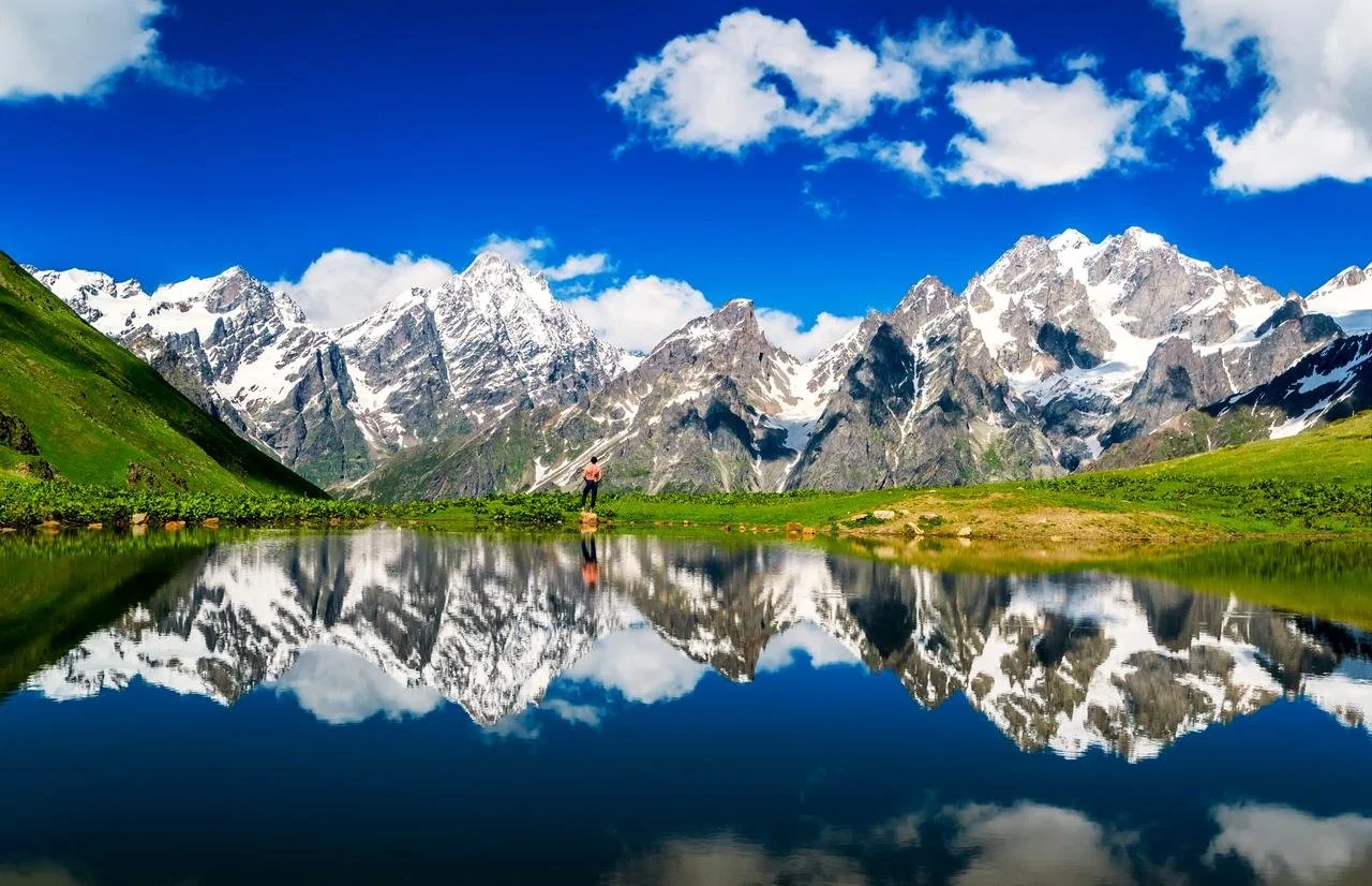 A person standing near a body of water with snow-capped mountains and a blue sky with clouds in the background, reflecting in the water.