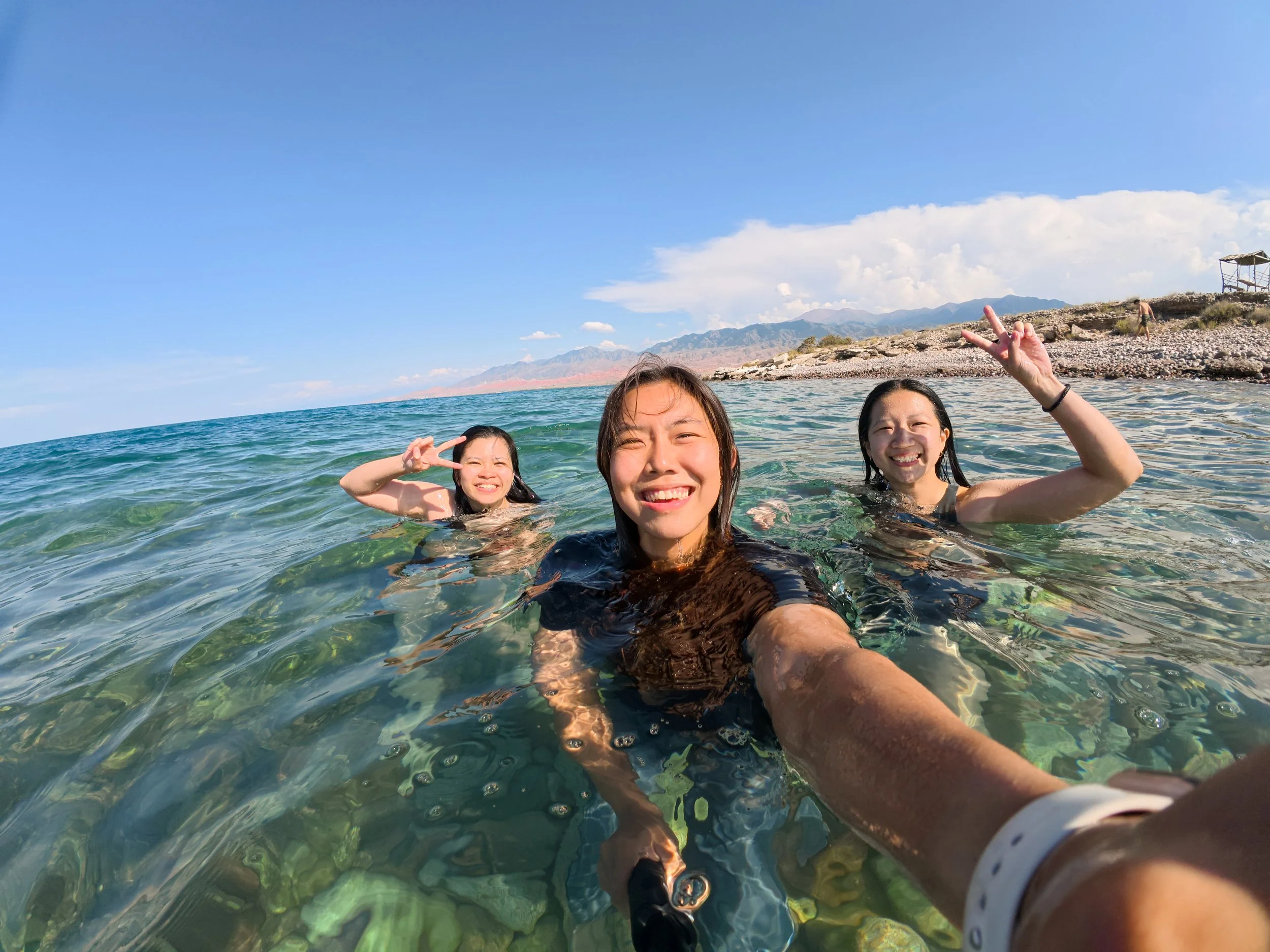 Three young women smiling and making peace signs while swimming in clear blue water at the beach on a sunny day.