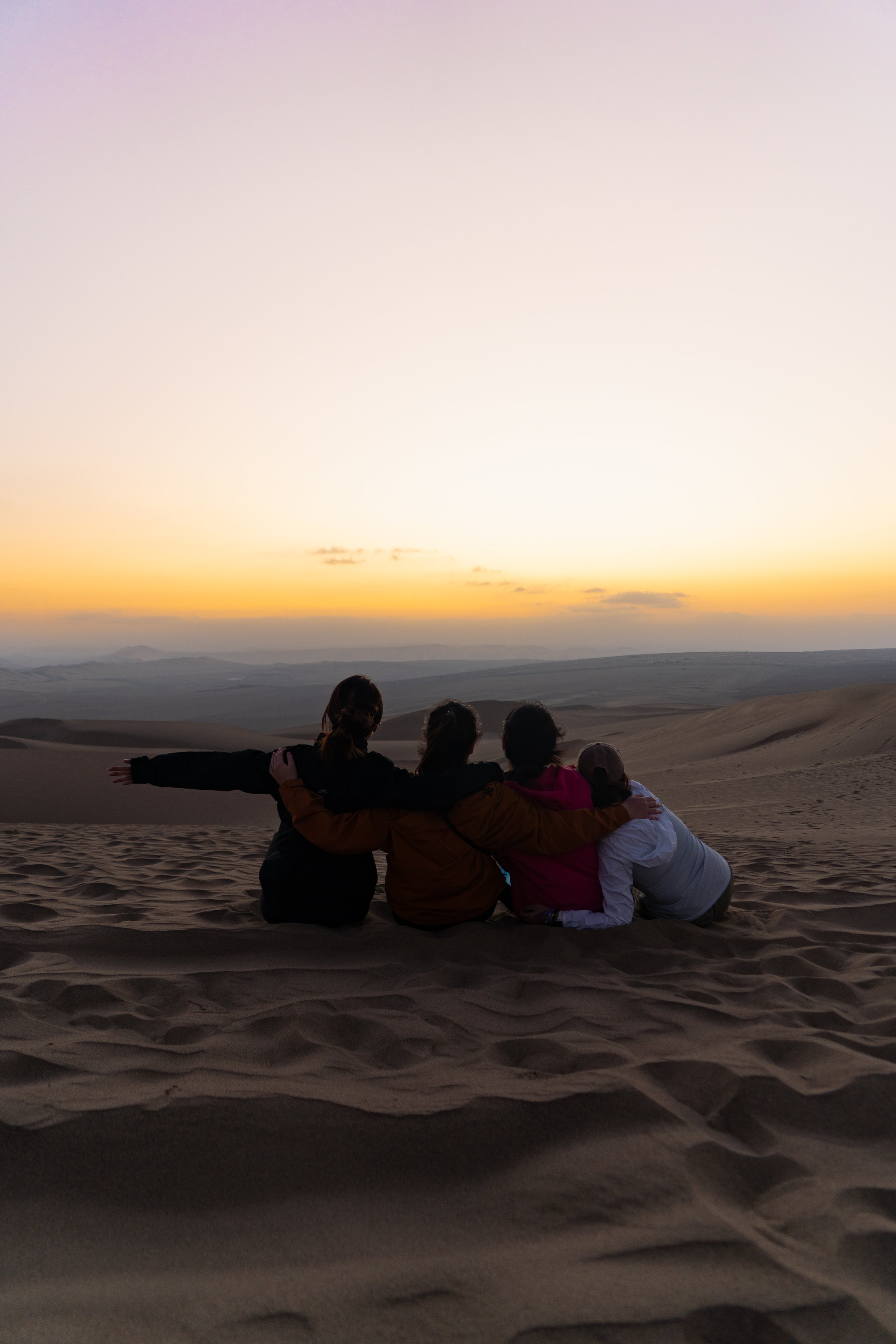 Peru, Huacachina. Four people sitting on sand dunes watching a sunset in a desert, with arms around each other.