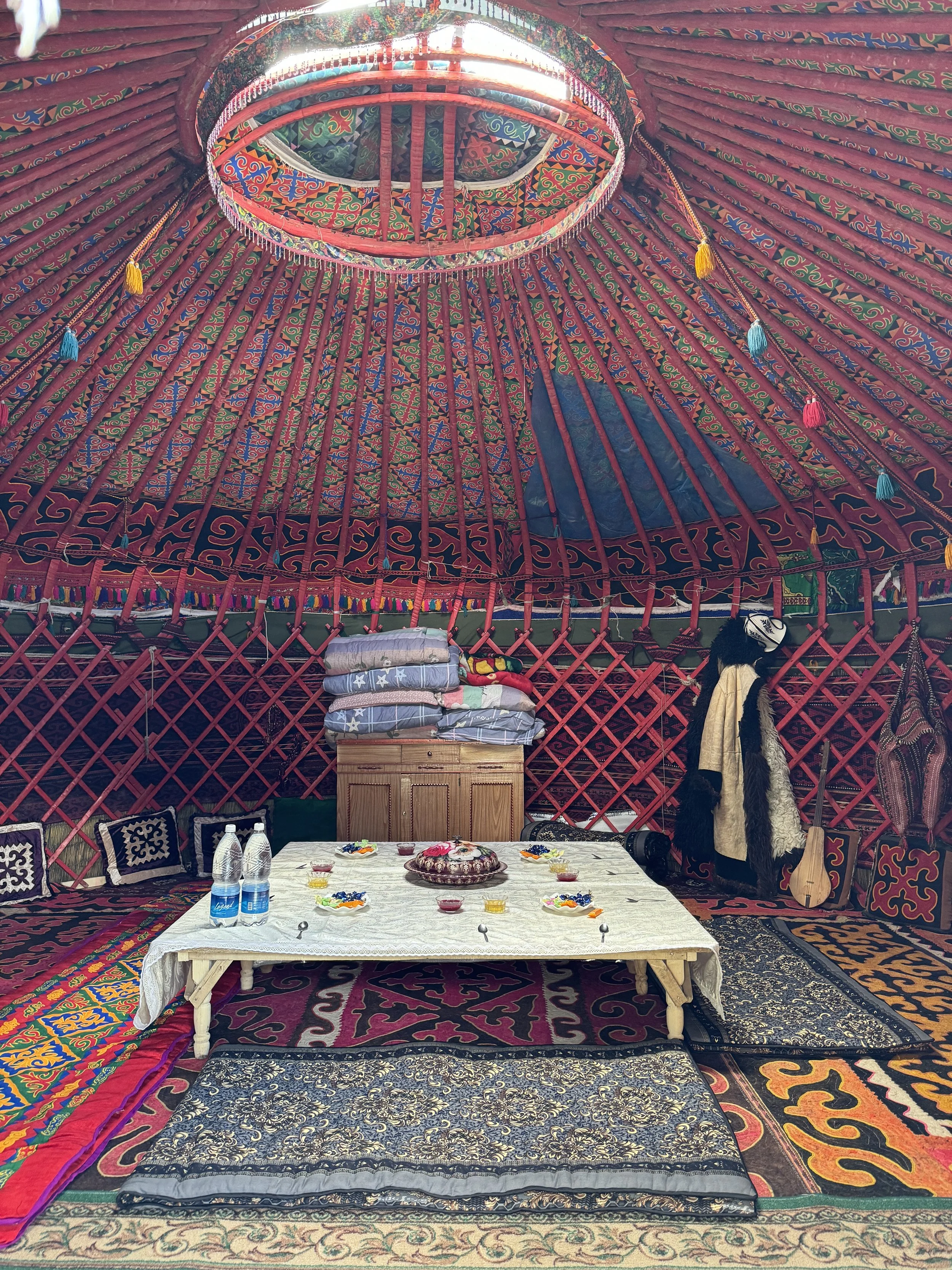 Inside of a colorful yurt decorated with patterned fabrics and carpets, with a low table set with plates of food and drinks, and traditional clothing hanging inside.