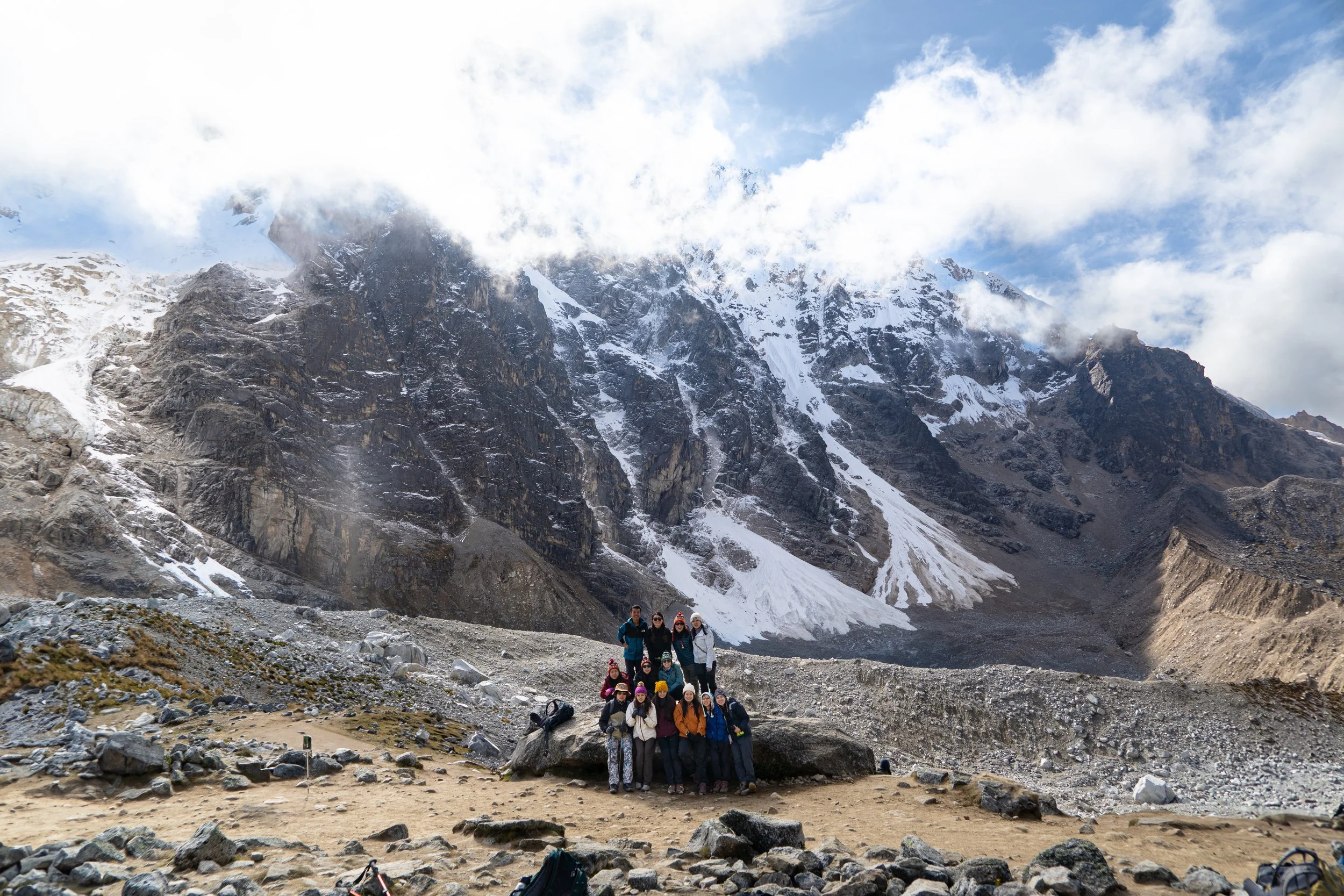 Group of hikers standing on rocky terrain at a mountain base, with snow-covered peaks and clouds in the background.