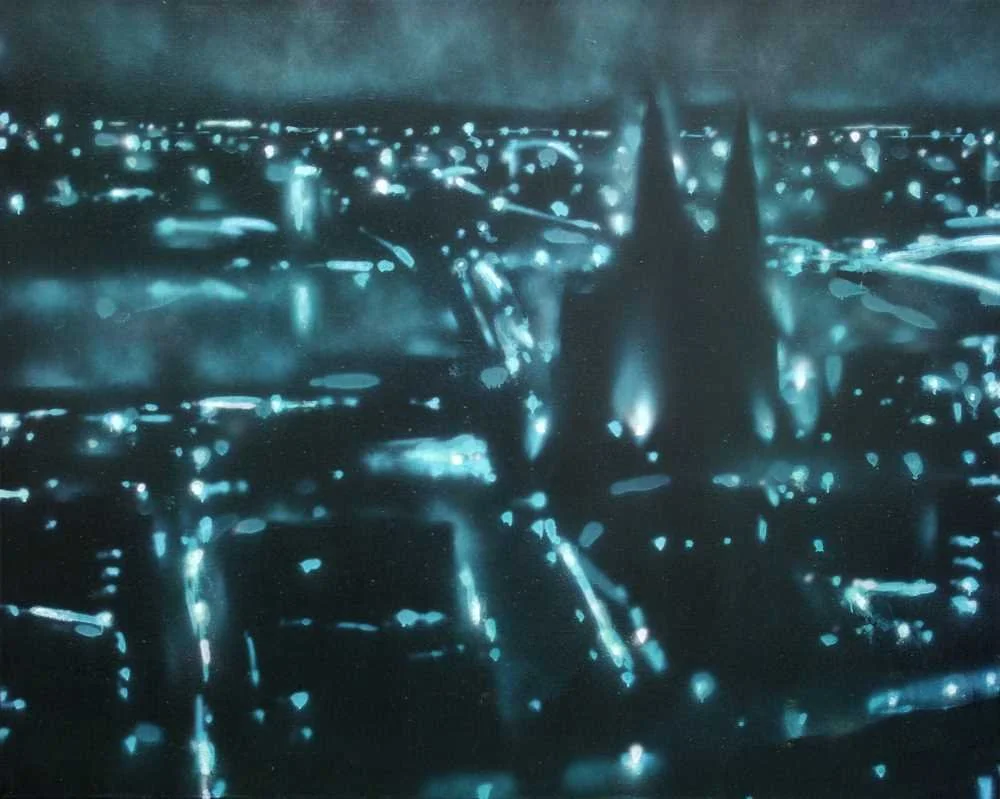 Ein unscharfes Bild von einer Stadt bei Nacht mit leuchtenden Lichtern und einem dunklen Himmel. Im Vordergrund ist eine Straße mit reflektierenden Oberflächen zu sehen.