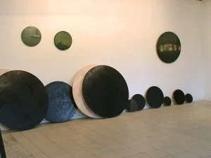 Verschiedene schwarze und graue runde Objekte in unterschiedlicher Größe, an einer weißen Wand, einige am Boden aufgereiht, einige an der Wand montiert.