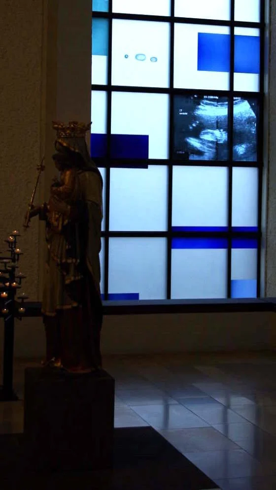 Eine Statue eines heiligen Mannes mit Krone, die neben einem Kerzenständer steht, im Vordergrund. Hinter ihm ist ein Fenster mit abstrakten blauen und weißen Mustern, durch das ein Ultraschallbild sichtbar ist.