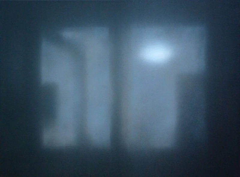 Das Bild zeigt eine dunkle, ruhige Innenraumszene mit einem Fenster, durch das Licht auf eine Wand fällt.