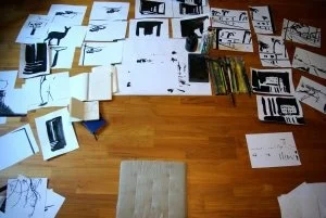 Viele verstreute Papiere mit Text und Bildern auf einem Holzboden, ein Sitzkissen im Vordergrund.