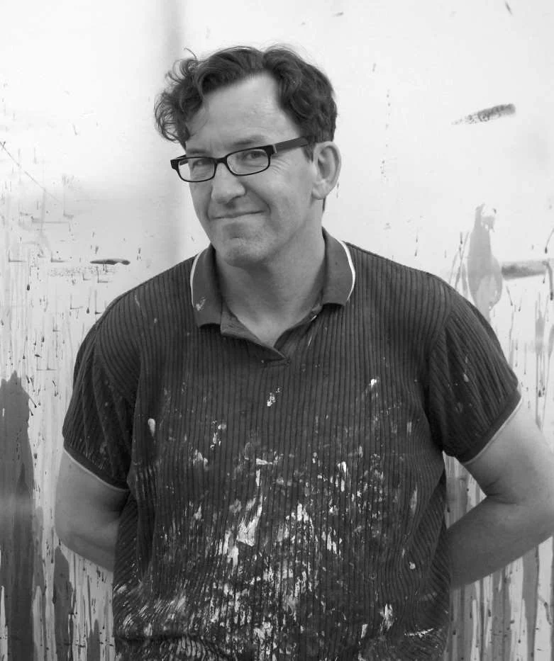 Mann mit Brille, lockigen Haaren, trägt ein dunkles, gestreiftes T-Shirt mit Farbklecksen, steht vor einer whiteboardartigen Wand mit Farbspritzern.