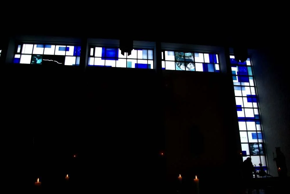 Innenraum einer Kirche mit Buntglasfenstern in Blau- und Weißtönen, dunkler Raum, paar Kerzen und zwei Menschen sind zu sehen.