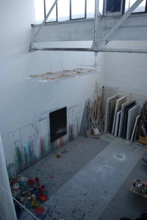 Ein Kunststudio mit einer großen, hängen Installation in der Luft, Malfarben an der Wand, und auf dem Boden künstlerische Materialien und Werkzeuge.
