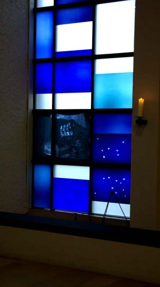 Blaue und weiße geometrische Fensterfront in einem Gebäude, mit einer Kerze an der rechten Seite