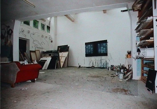 Ein Raum mit einem Fernseher an der Wand, eine rote Couch, mehrere Rahmenbilder und Bücherregale an den Wänden, sowie Mal- und Zeichenutensilien auf dem Boden und in Regalen.