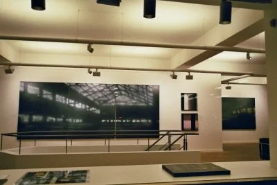 Ein Ausstellungsraum mit mehreren großformatigen Fotografien an weißen Wänden und Deckenlampen.