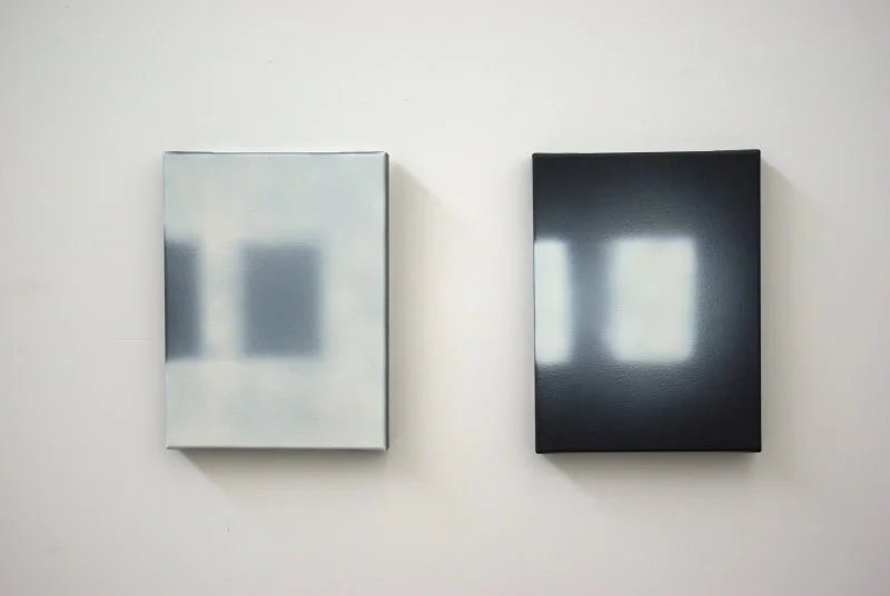 Zwei minimalistische abstrakte Gemälde, das linke ist in Silber- und Grautönen, das rechte in Schwarz gehalten, hängen an einer weißen Wand.