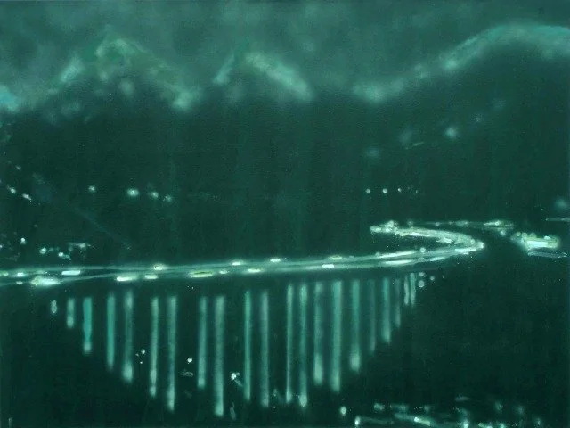 Nachtaufnahme einer Brücke über ein Wasser mit Bergen im Hintergrund, beleuchtet mit Straßenlaternen.