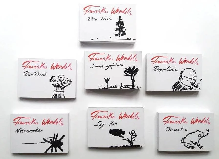 Sechs kleine Bücher mit schwarzen Illustrationen und roten handgeschriebenen Titeln von Franciscus Wendels, die verschiedene Themen wie den Trieb, den Durst, Sonntagfahreter, Doggen, Netzwerke, Sig-Kub und Prinzesskuss zeigen, sind auf weißem Hintergrund angeordnet.