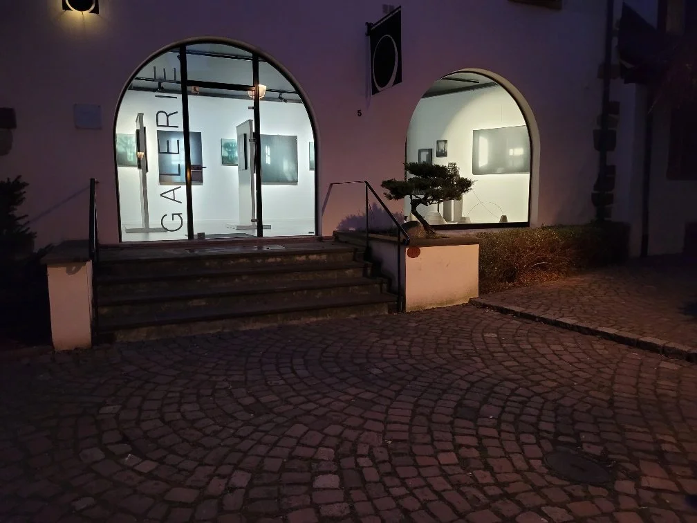 Kunstgalerie mit großen, runden Fenster, beleuchtet bei Nacht, mit Treppe davor, Außenpflanzen, beschildert mit 'GALER'