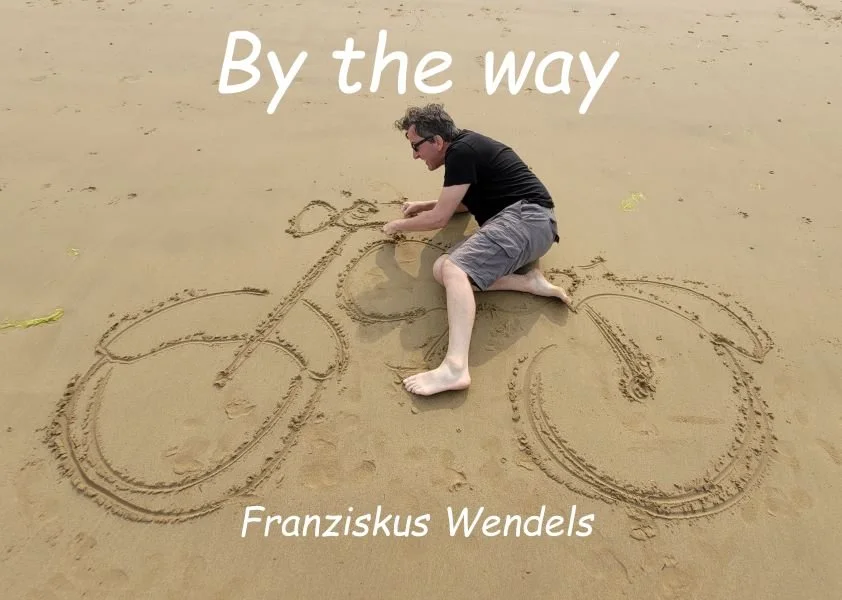 Ein Mann zeichnet ein Fahrrad in den Sand am Strand, mit Text "By the way" und "Franziskus Wendels" darüber und darunter.