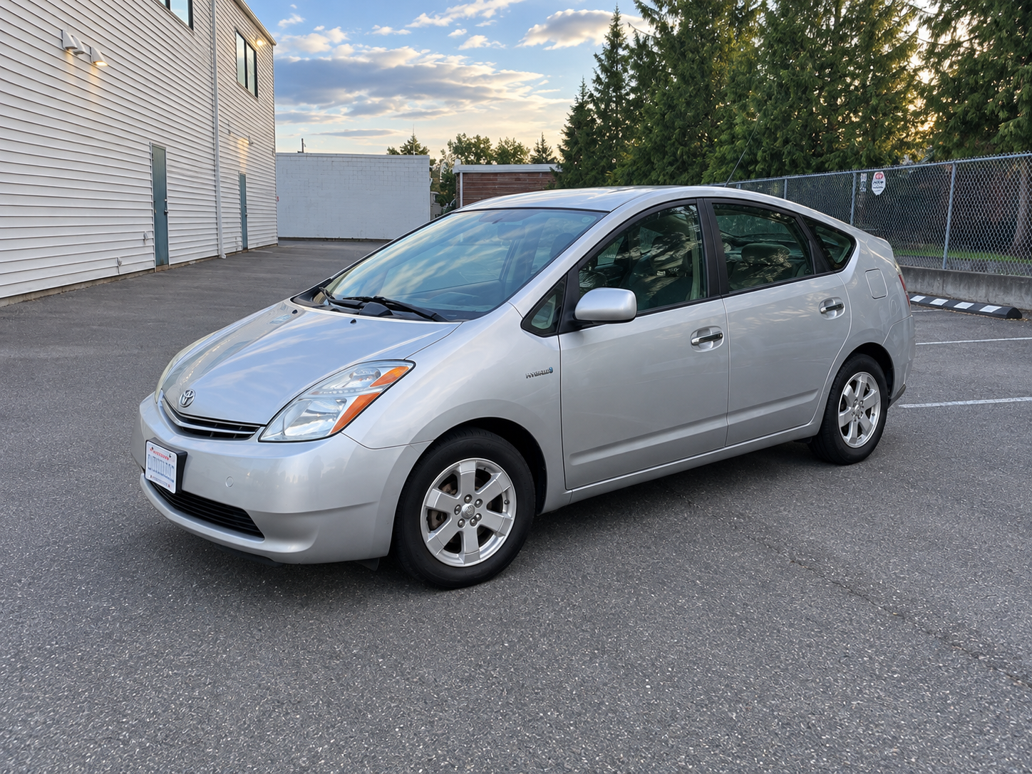 Silver Toyota Prius hybrid hatchback parked in an outdoor lot beside a building and chain-link fence.