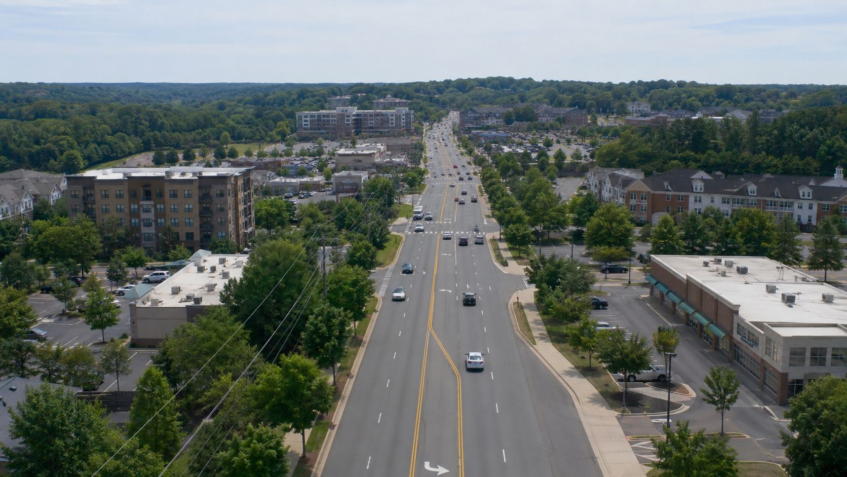 Aerial view of a street in a suburban area with cars driving along the road, surrounded by trees, commercial buildings, and apartment complexes, with a backdrop of green hills.
