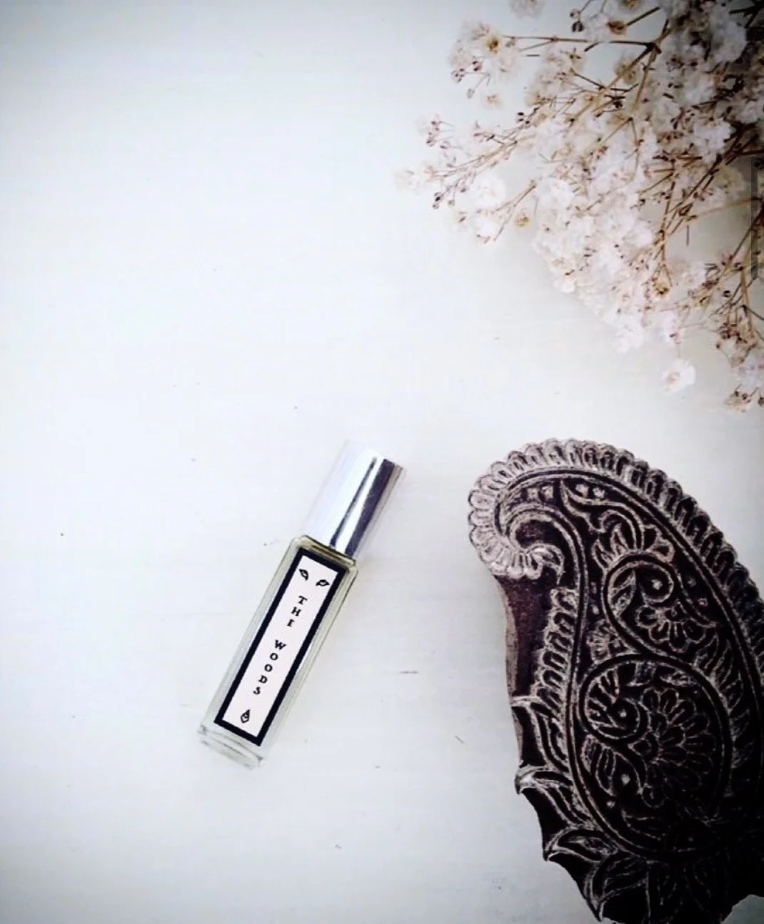 A natural perfume bottle labeled "The Woods" on a white surface, with a dark patterned textile and a cluster of white flowers nearby.