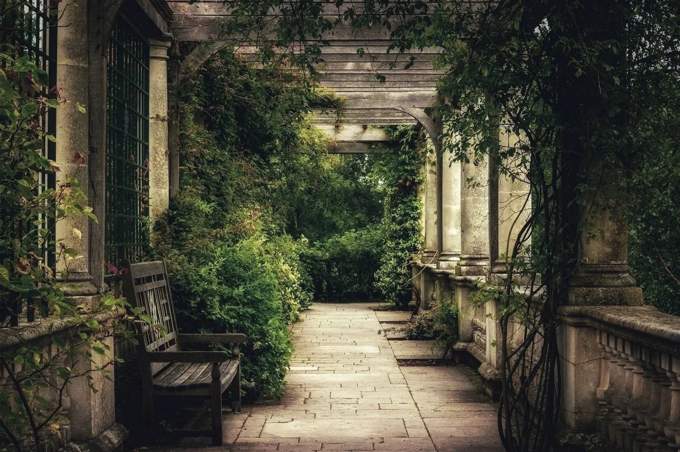 A stone pathway through a lush garden corridor with an arched wooden pergola, surrounded by greenery and stone columns.
