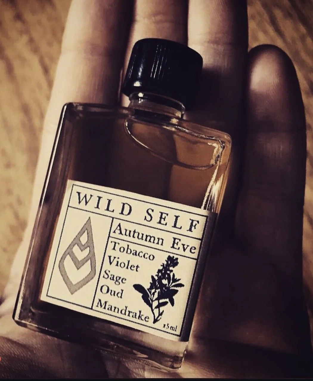 A bottle of cologne labeled 'Wild Self' with scents such as autumn eve, tobacco, violet, sage, oud, and mandrake, held in a person's hand.