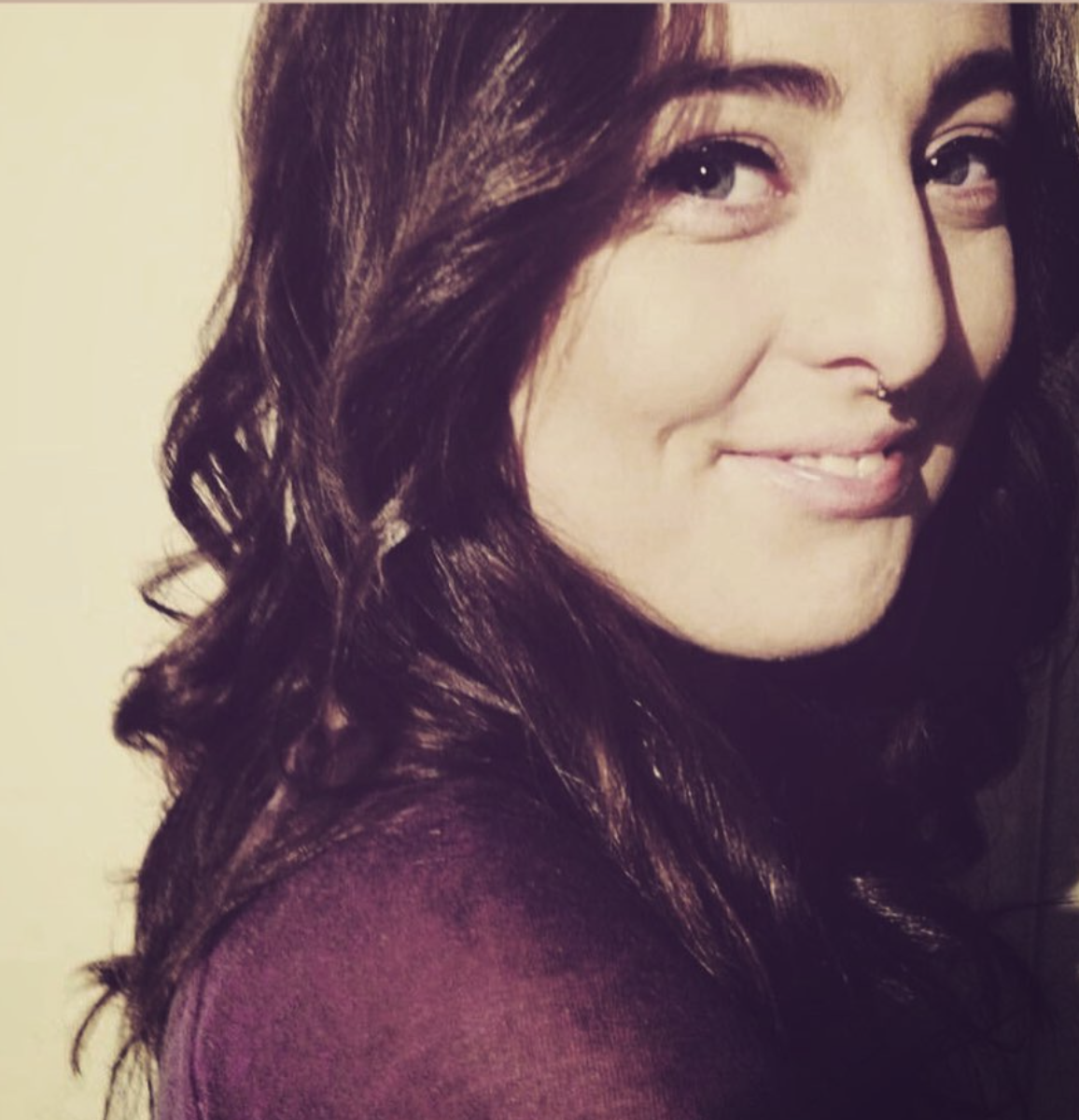 Close-up of a woman with long, wavy dark hair, smiling, with a septum piercing, wearing a purple top.